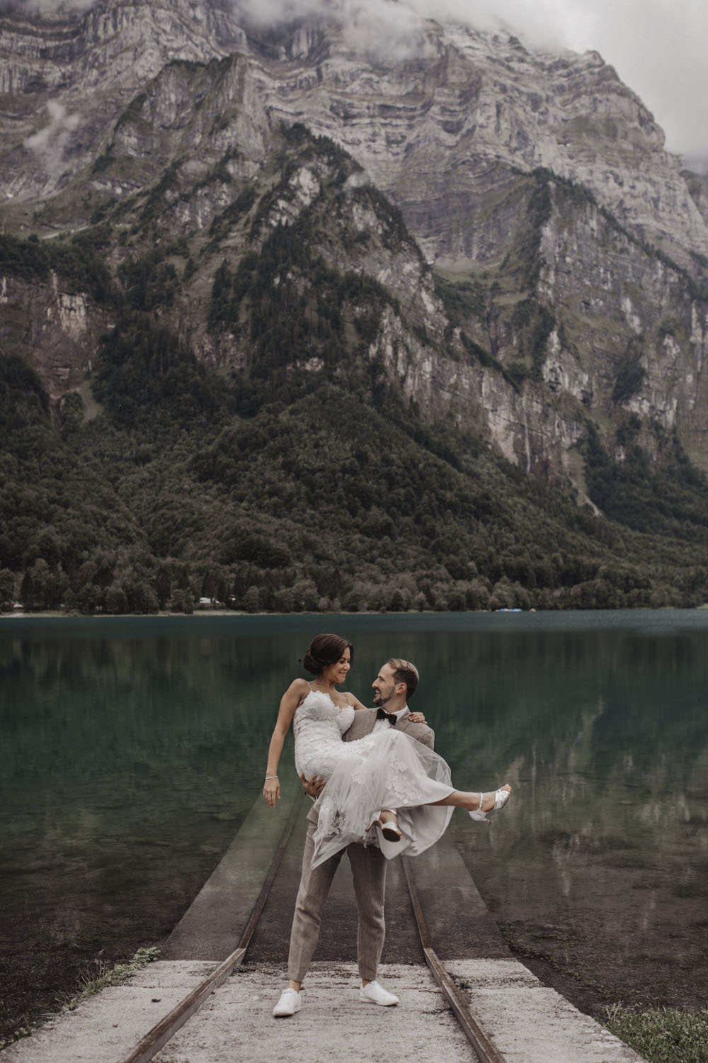 Bräutigam trägt Braut lachend am Seeufer