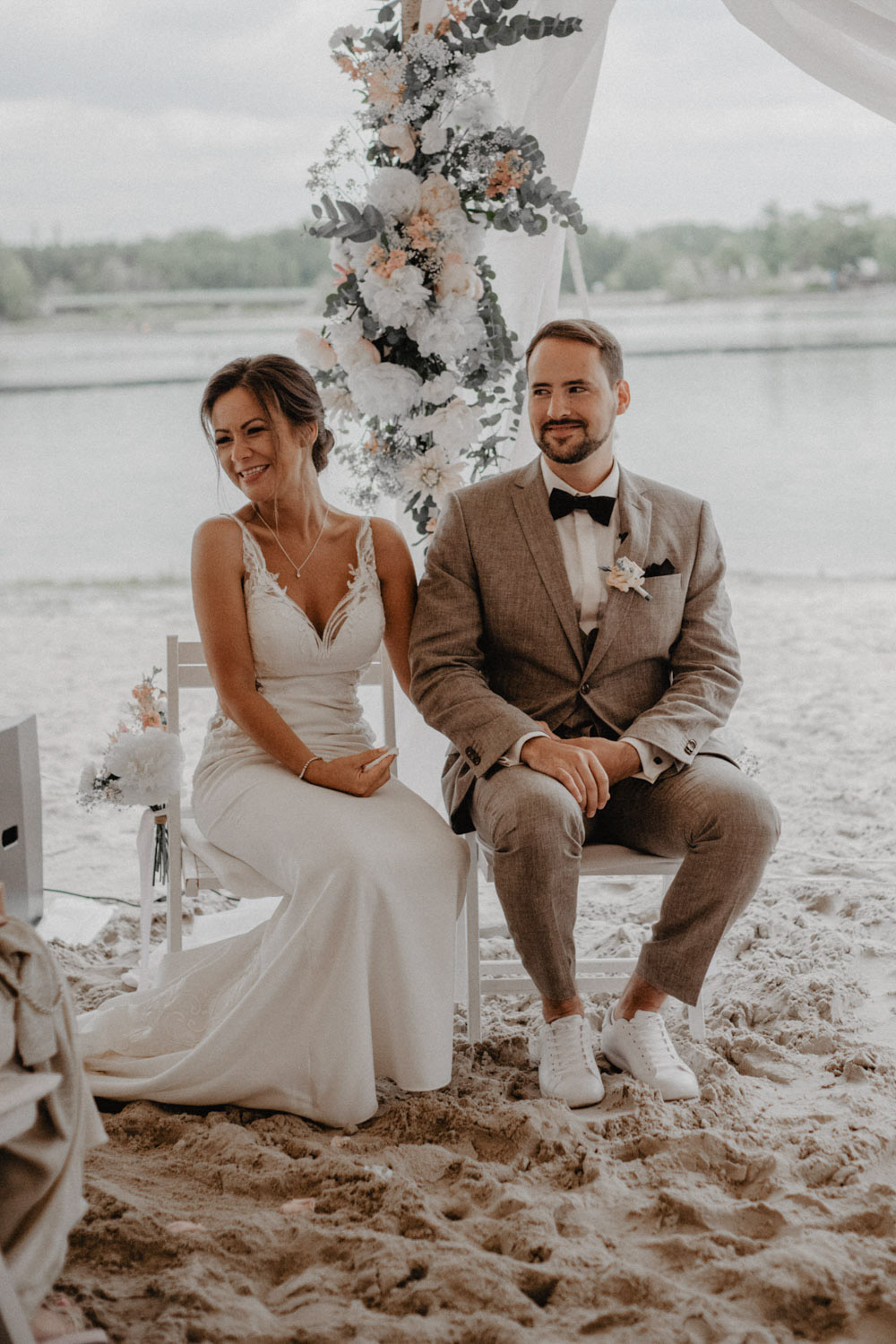 Brautpaar sitzt lächelnd vor dem floralen Traubogen am See