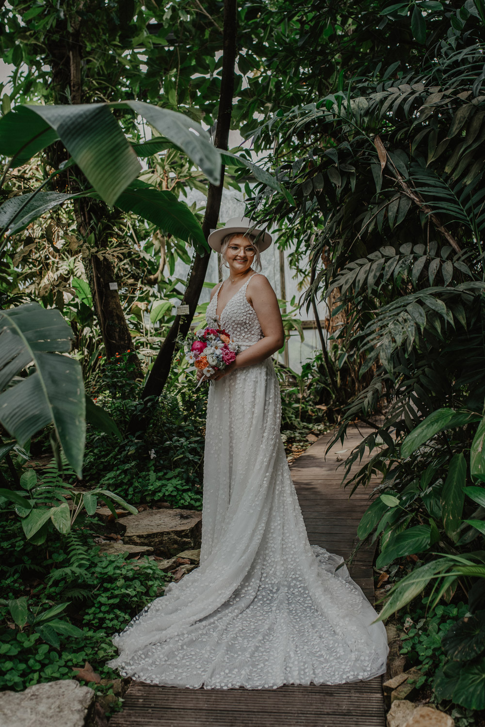 Braut mit Hut und Blumenstrauß steht im Regenwaldhaus
