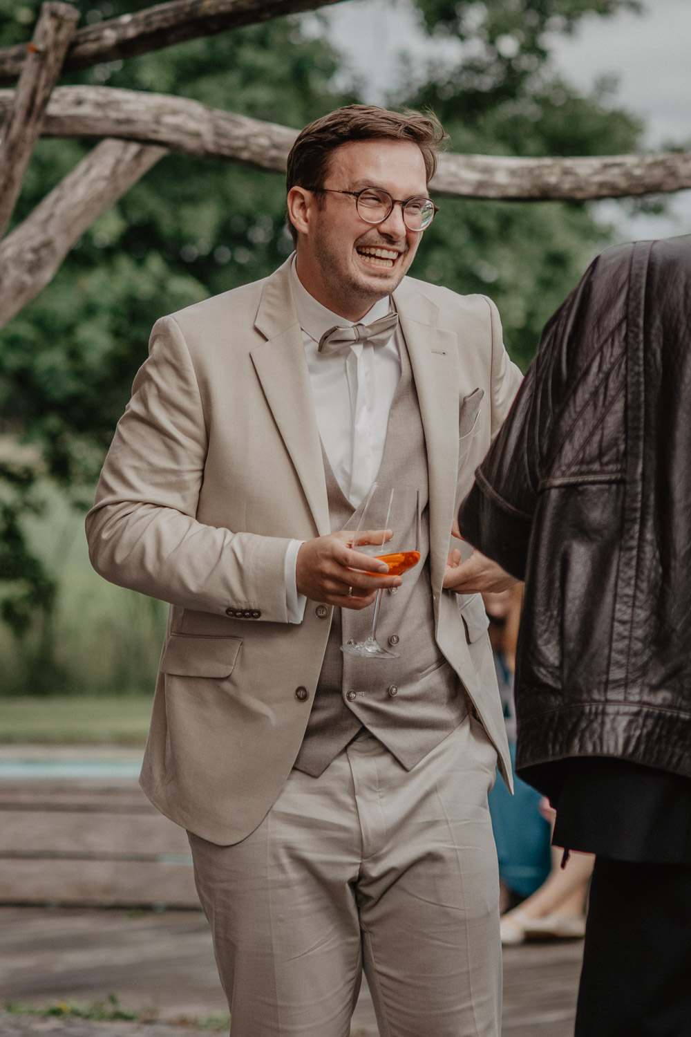 Bräutigam im beigen Anzug lacht mit Drink in der Hand
