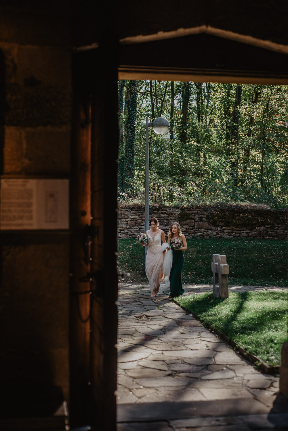 Braut kommt mit Brautjungfer über den Kirchhof zum Eingang.