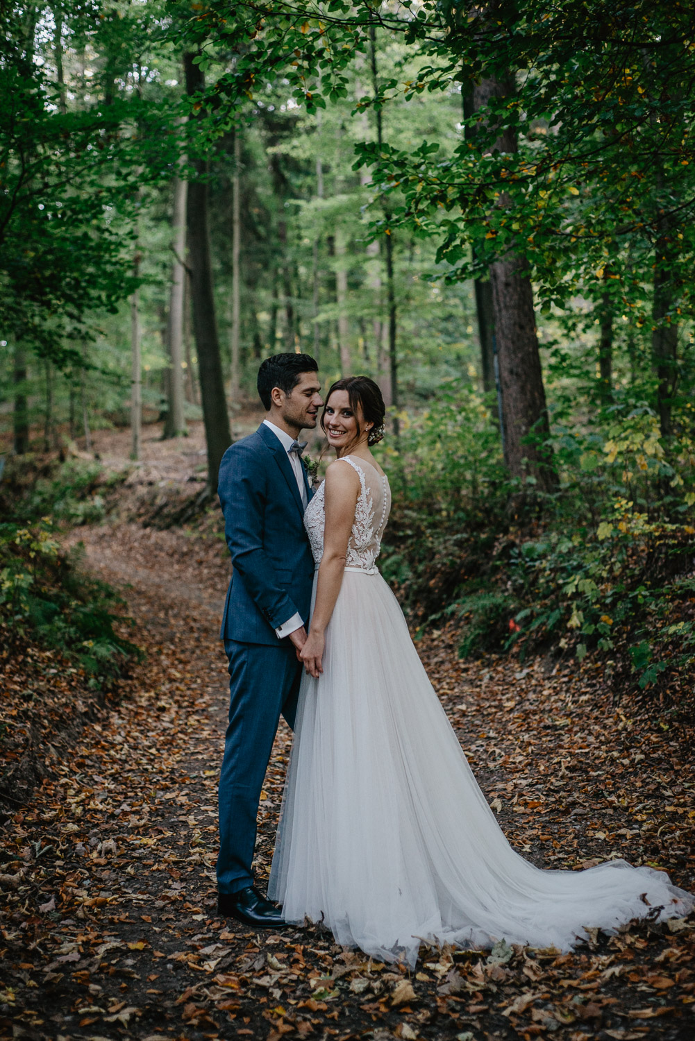 Brautpaar steht Hand in Hand im Wald und schaut in die Kamera.