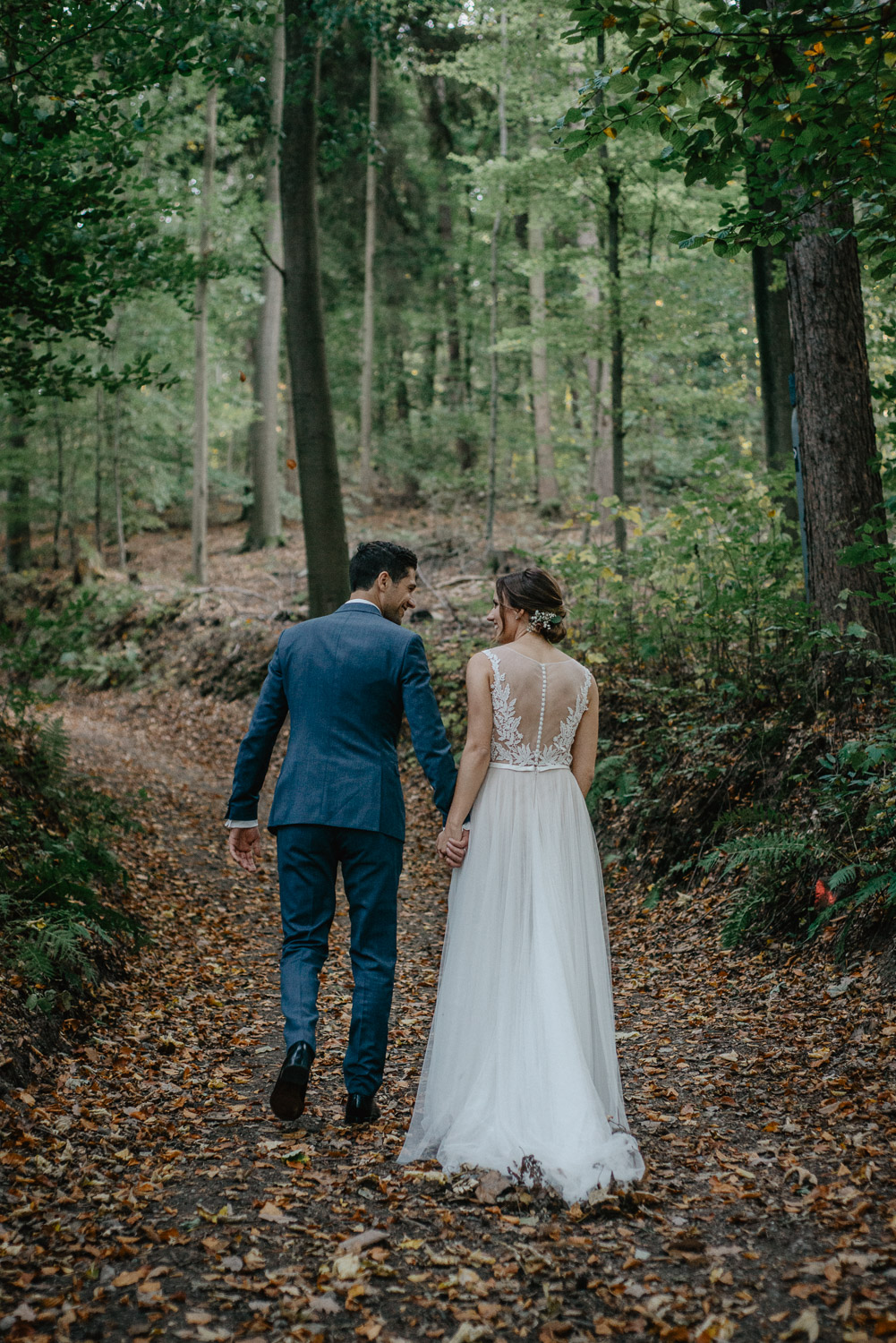 Braut und Bräutigam gehen Hand in Hand einen Waldweg entlang.