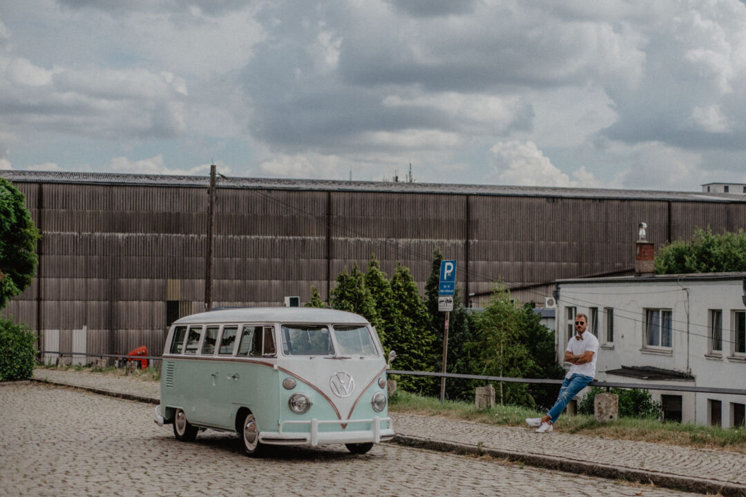 Mintfarbener VW Bulli steht vor industrieller Kulisse