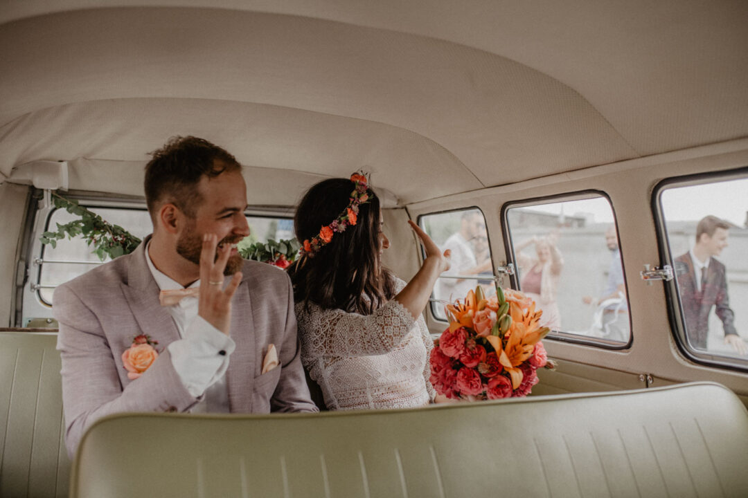 Brautpaar winkt aus dem VW Bulli nach der Trauung