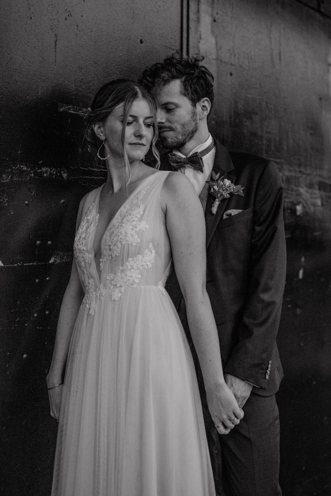 Bräutigam lehnt sich liebevoll an Braut vor dunkler Wand