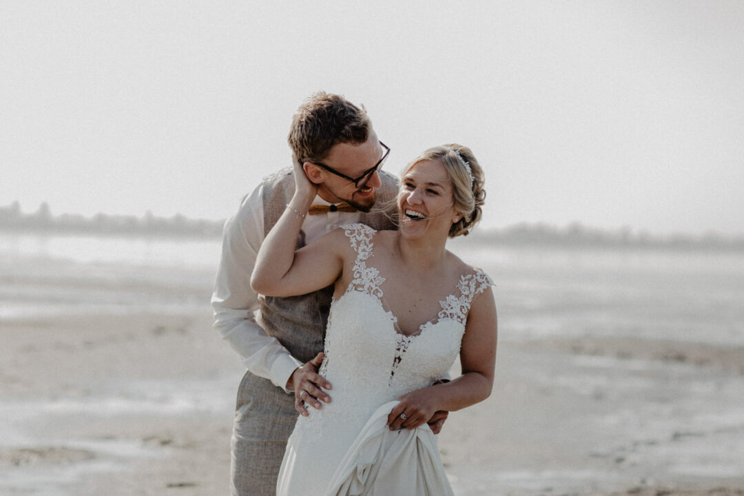 Bräutigam umarmt die Braut von hinten am Strand