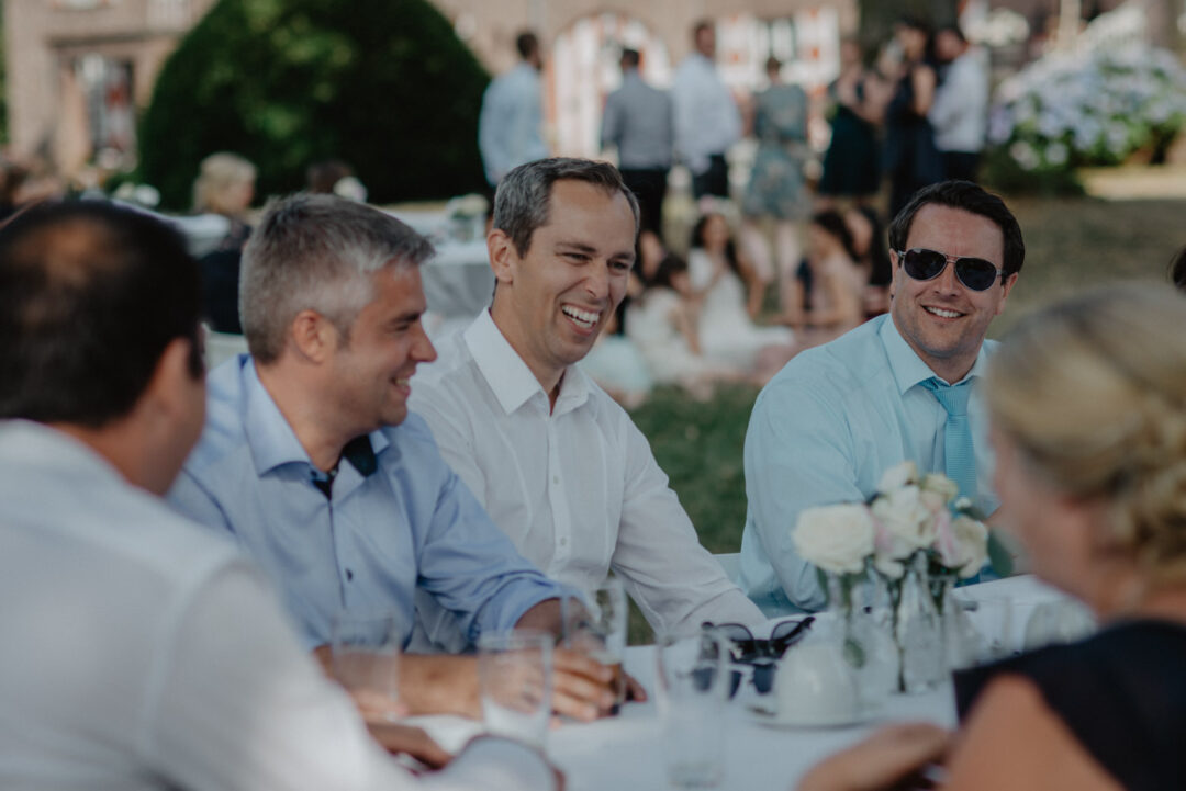 Maenner lachen gemeinsam am Tisch auf der Hochzeit