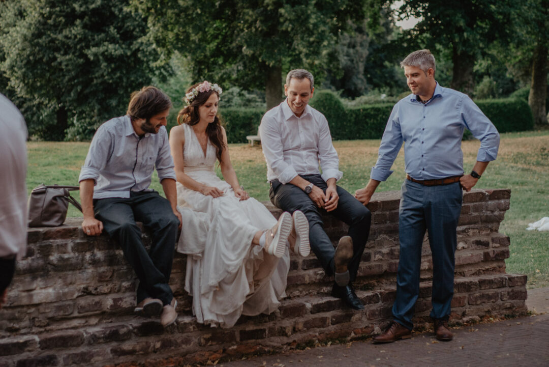 Braut sitzt mit Freunden auf Mauer bei Hochzeit