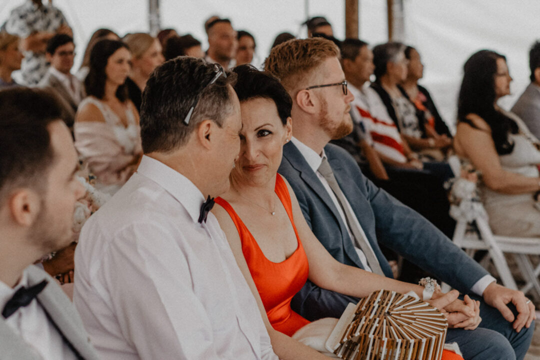 Gäste sitzen gespannt unter Zeltdach bei Strandhochzeit am See