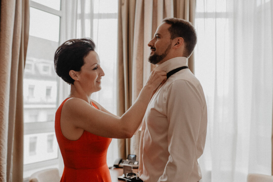 Frau hilft Bräutigam beim Binden der Fliege im Hotelzimmer