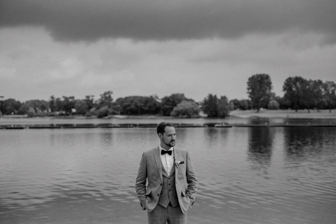 Bräutigam steht alleine vor ruhigem Wasser am Fühlinger See