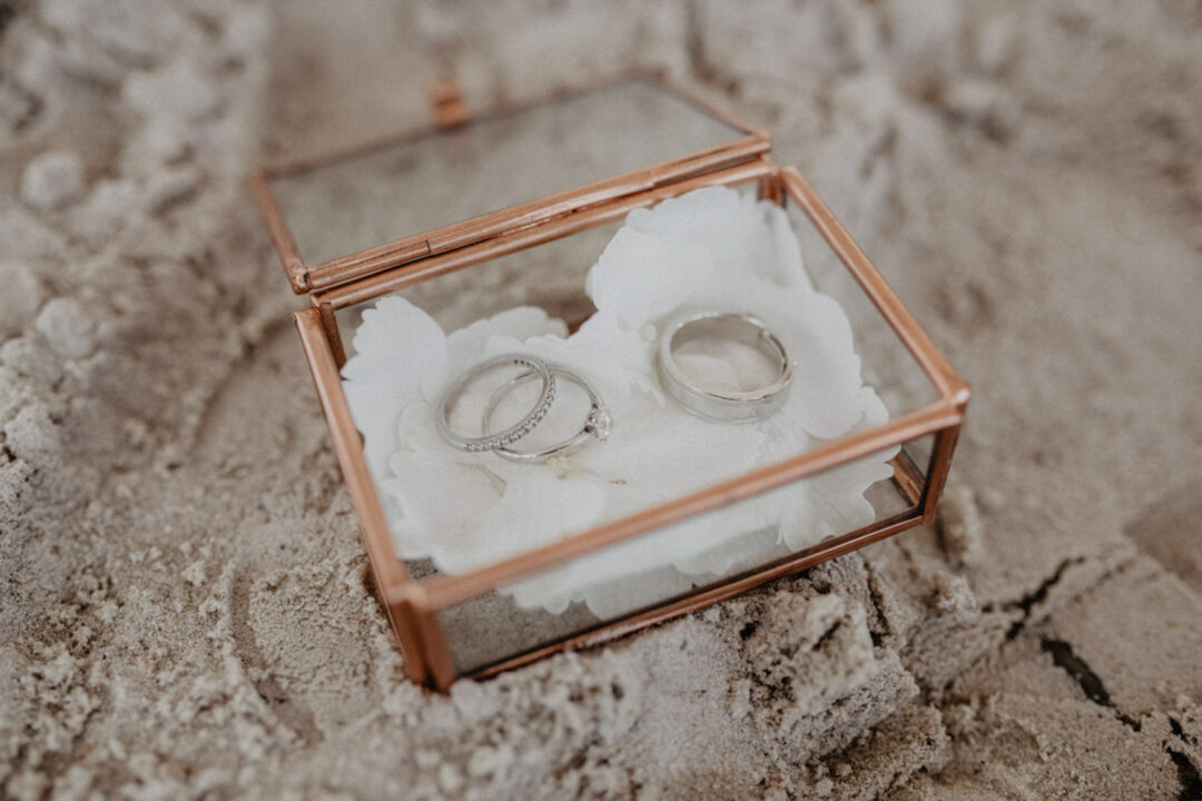 Silberne Eheringe in Glasbox mit Blüten auf Sand