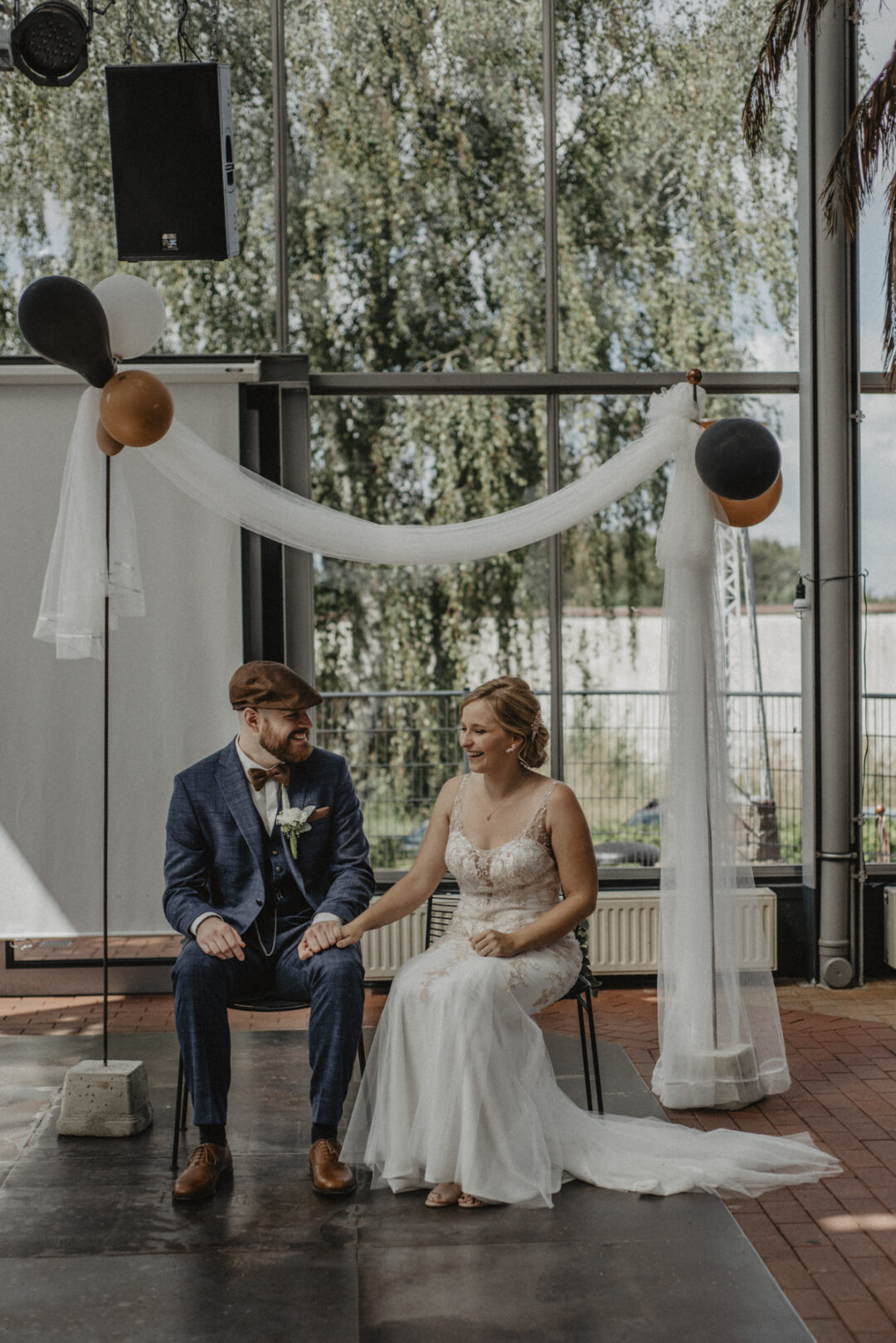 Brautpaar sitzt Hand in Hand auf der Bühne unter einem Traubogen mit Ballons