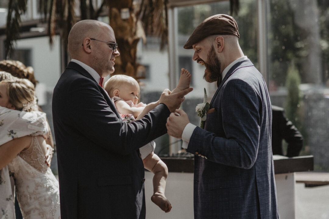Bräutigam begrüßt lachend ein Baby, das von einem Gast auf dem Arm gehalten wird