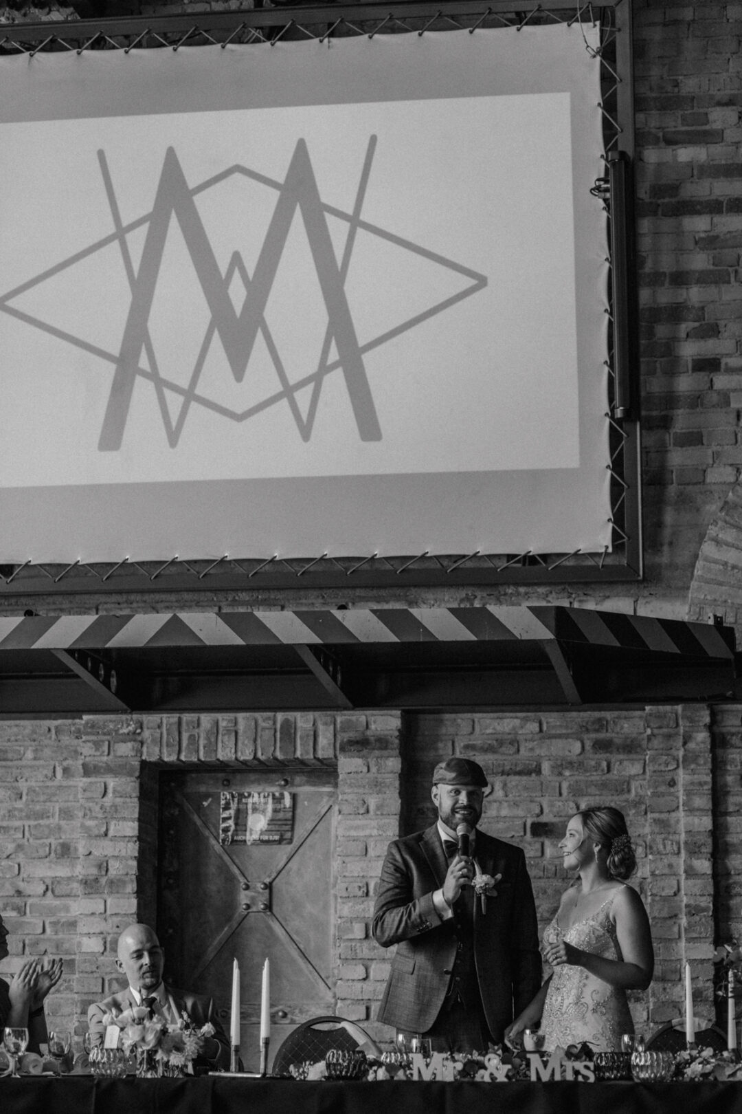 Bräutigam hält Rede neben der Braut unter einer Projektion mit Logo