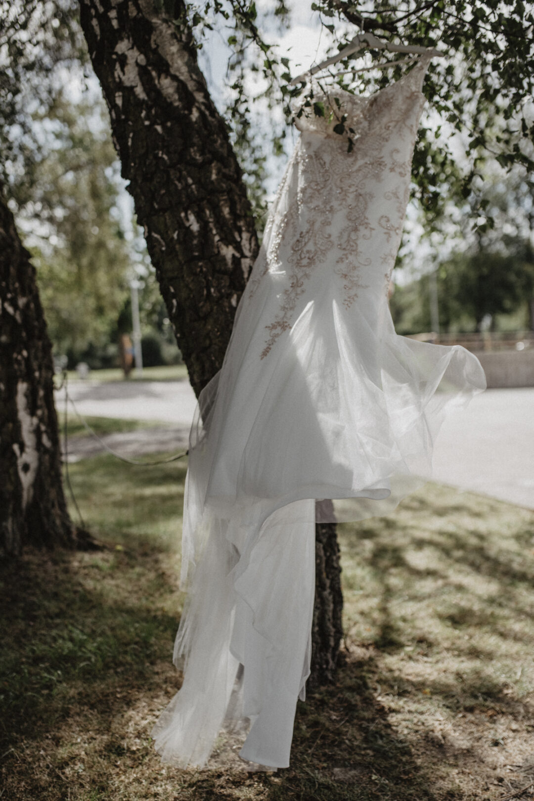 Brautkleid weht im Wind – Detailaufnahme vor rustikalem Baum im industriellen Ambiente