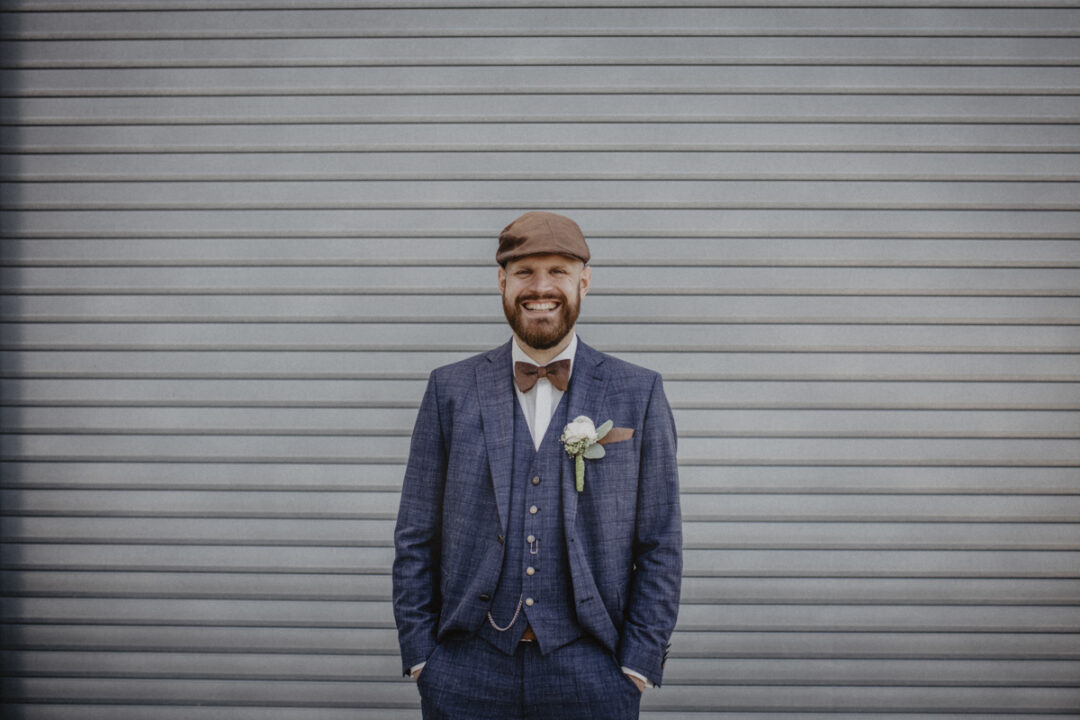Bräutigam in blauem Anzug und Schiebermütze steht entspannt vor einem grauen Garagentor
