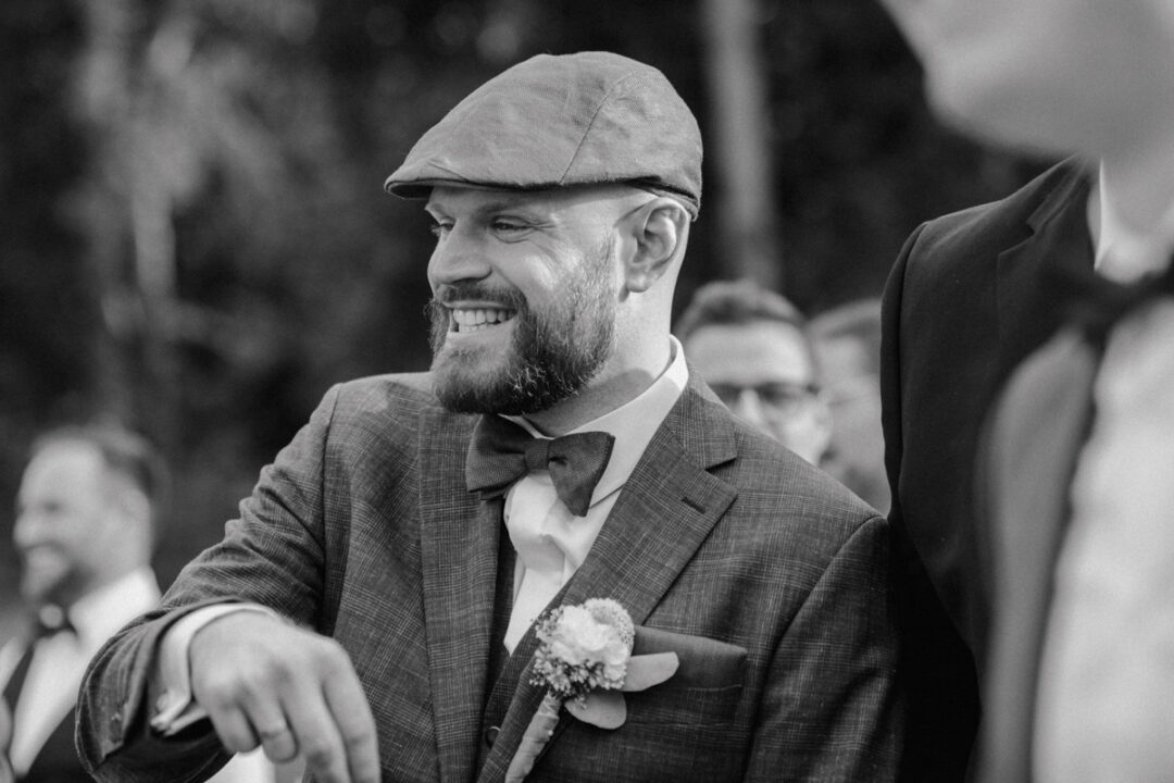 Schwarz-weiß-Porträt eines lächelnden Bräutigams mit Fliege und Ansteckblume