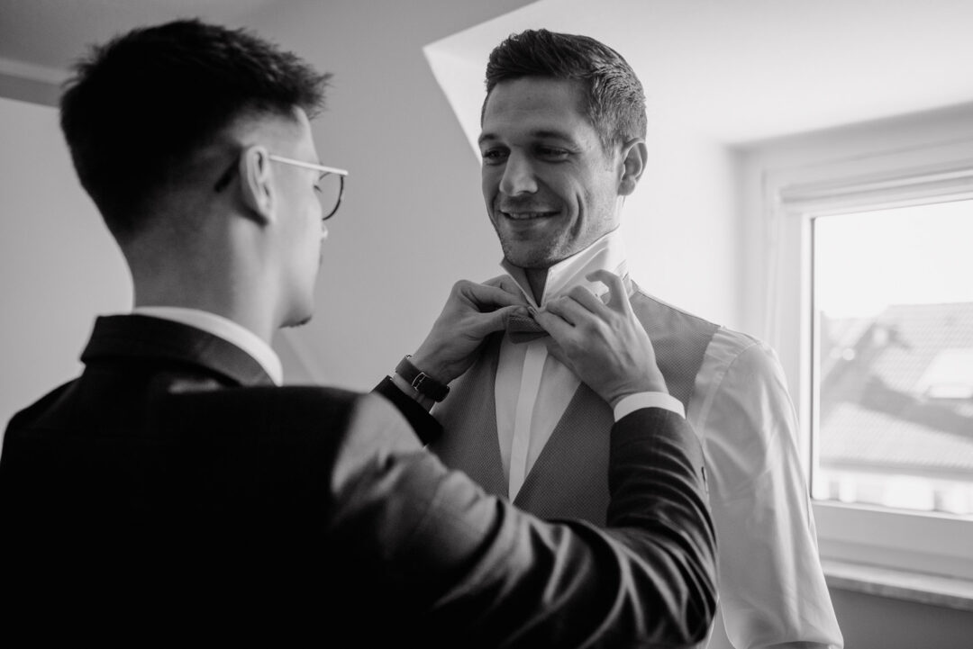 Bräutigam bekommt Hilfe beim Anlegen der Fliege, Schwarz-Weiß Aufnahme.