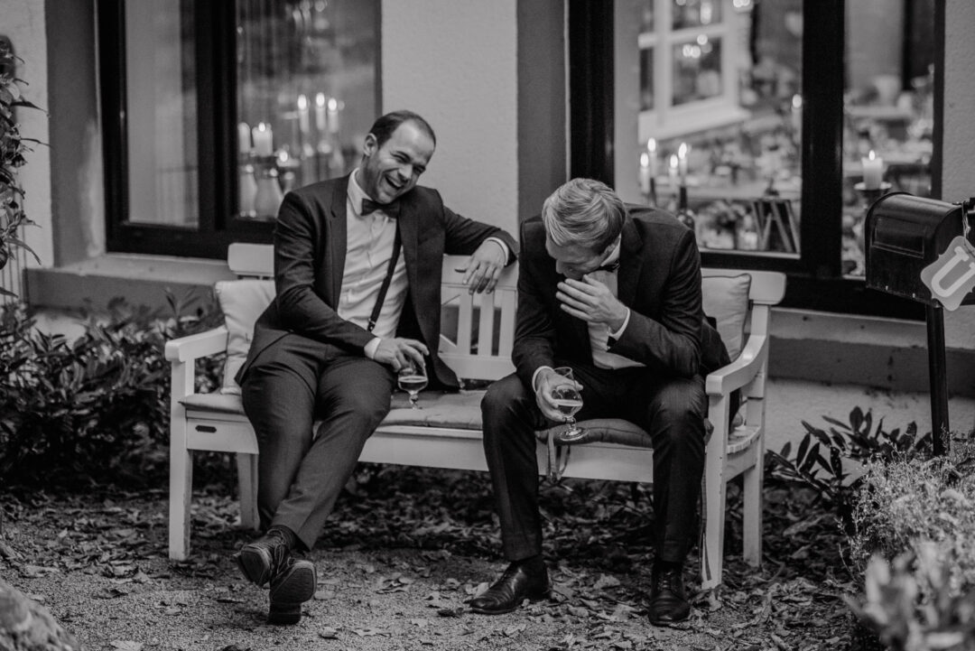 Zwei männliche Gäste lachen herzlich auf einer Bank vor dem Wirtshaus, Schwarz-Weiß.