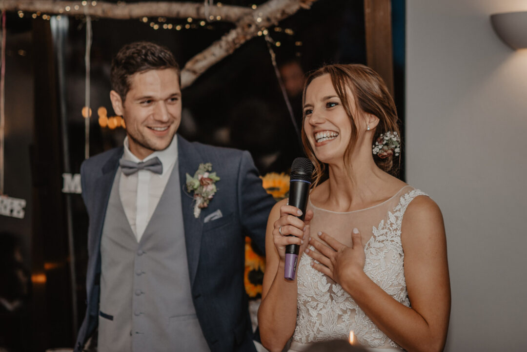 Braut spricht mit Mikrofon, Bräutigam steht lachend daneben.