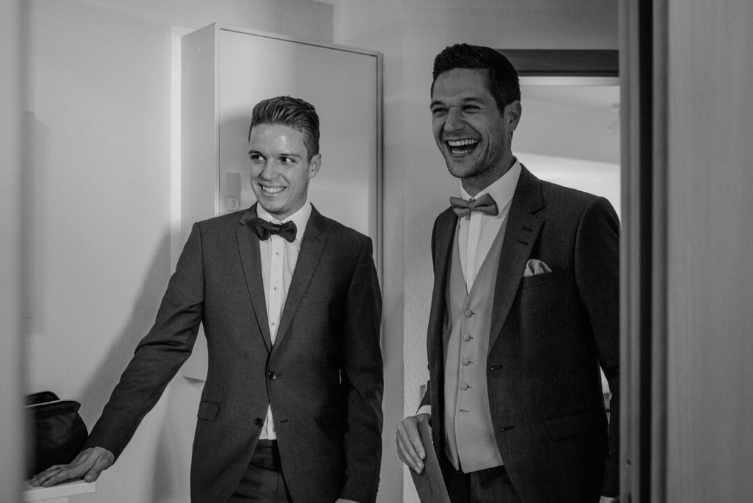 Bräutigam lacht gemeinsam mit Trauzeugen beim Getting Ready, Schwarz-Weiß.