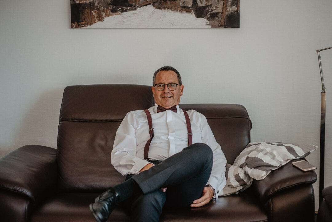 Vater der Braut sitzt entspannt in festlicher Kleidung auf dem Sofa.