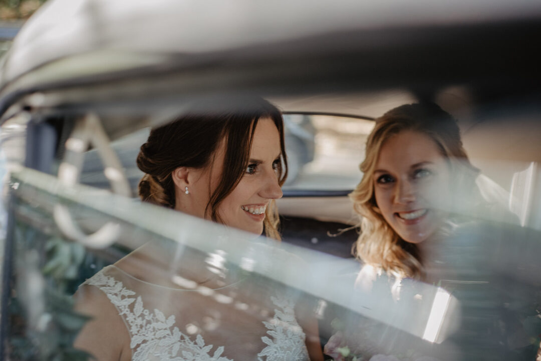 Braut sitzt mit Brautjungfer lächelnd im Auto vor der Kirche.