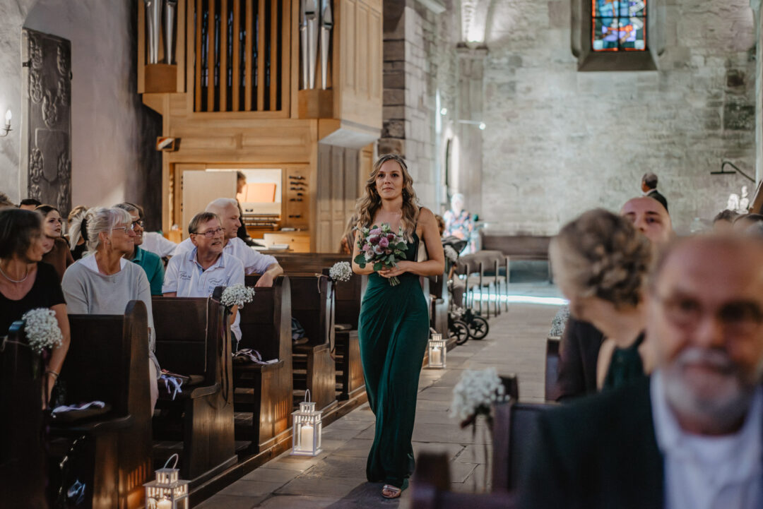 Brautjungfer schreitet mit Blumenstrauß den Mittelgang der Kirche entlang.