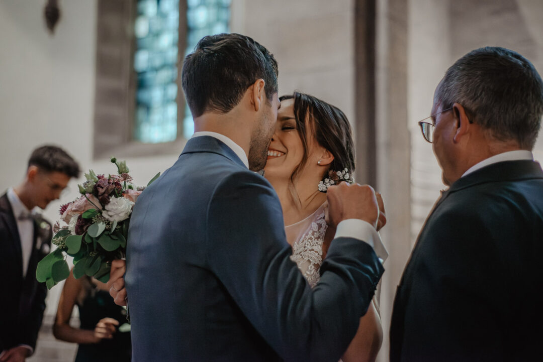 Bräutigam begrüßt seine Braut mit einem Kuss am Altar.
