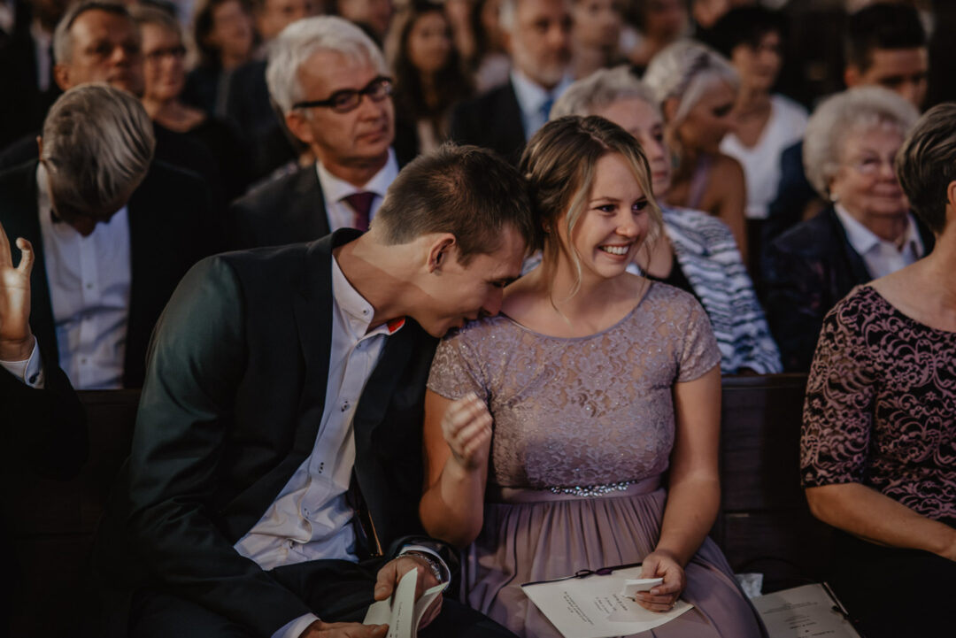 Junge Gäste lachen gemeinsam in der Kirchenbank während der Trauung.