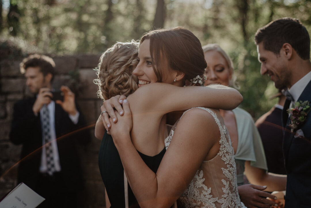 Braut umarmt eine Freundin herzlich draußen vor der Kirche.