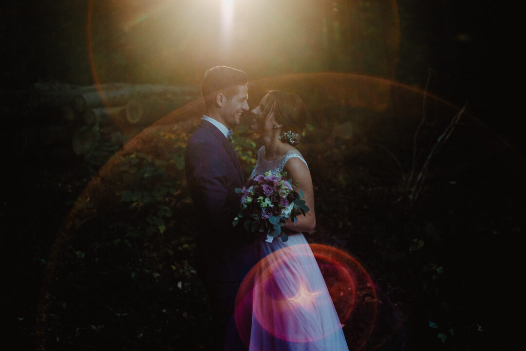 Brautpaar steht im Wald, Sonnenlicht strahlt durch die Bäume.