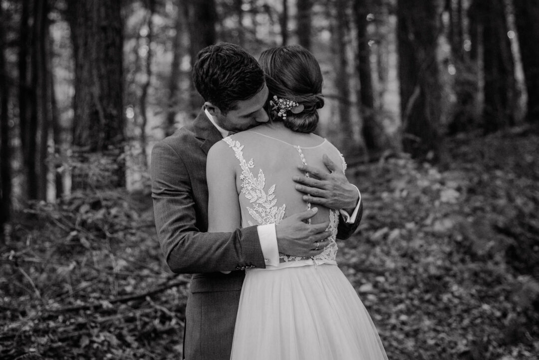 Bräutigam umarmt seine Braut liebevoll im Wald, Schwarz-Weiß Aufnahme.