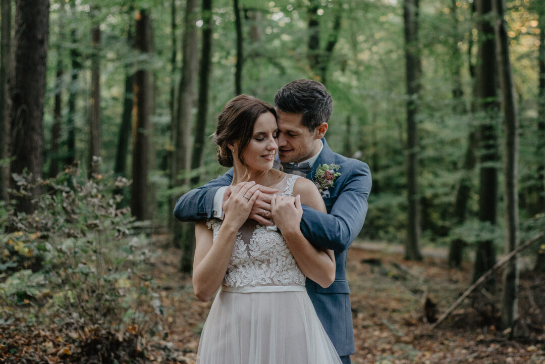 Bräutigam umarmt seine Braut liebevoll von hinten im Wald.