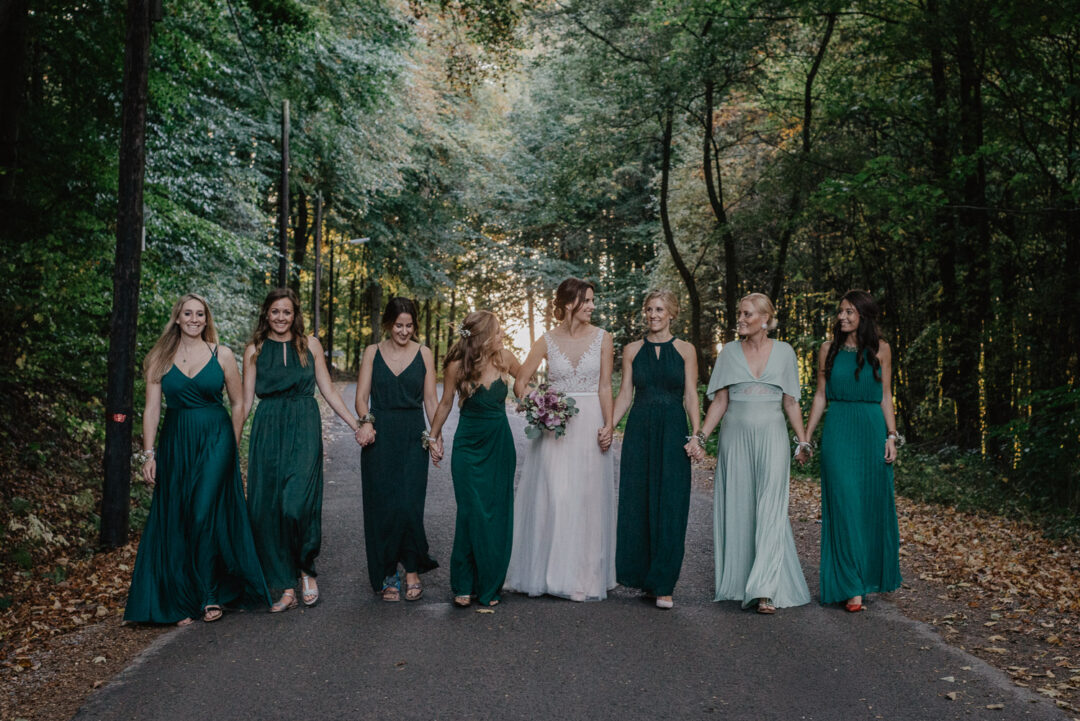 Braut geht mit ihren Brautjungfern Hand in Hand über eine Allee.